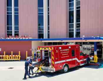 Picture of two male paramedics loading an empty stretcher into an ambulance in front of the emergency room of alhambra hospital medical center.
